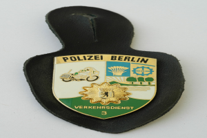 Ein schildförmiges Polizeisiegel mit 'Polizei Berlin' in schwarzer Fettschrift auf einem blauen Hintergrund mit einem weißen Stern und Rahmen, das auf einer Oberfläche ruht.