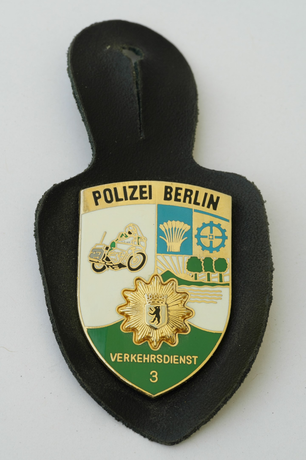 Ein schildförmiges Polizeisiegel mit 'Polizei Berlin' in schwarzer Fettschrift auf einem blauen Hintergrund mit einem weißen Stern und Rahmen, das auf einer Oberfläche ruht.
