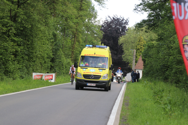 Ambulanz fährt auf einer Straße mit Radfahrern äquivalent, gesäumt von Gras, Bäumen, Häusern, Strommästen und einem klaren blauen Himmel.