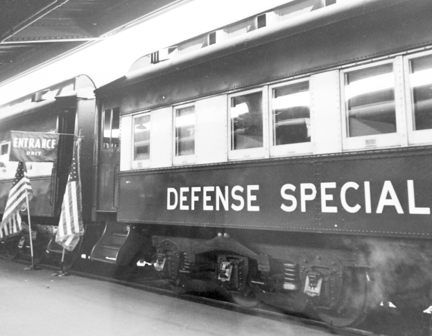 Schwarzes und weißes Foto eines Zuges mit der Aufschrift "Defense Special" auf den Schienen mit zwei Fahnen links und einer Decke oben.