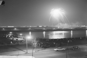 Ein Schwarz-Weiß-Foto von einer Menschenmenge, die Feuerwerk über Wasser betrachtet, mit Fahrzeugen, Laternenpfählen, Bäumen und Gebäuden im Hintergrund.