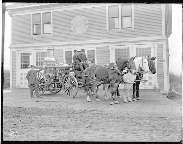 Schwarzes und weißes Foto von zwei Pferden, die einen Feuerwagen vor ein Gebäude ziehen, mit drei Personen im Wagen und zwei stehenden Personen in der Nähe, Bäume und klarer Himmel im Hintergrund.
