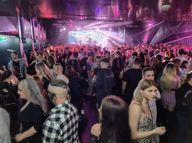 Eine große Menge steht in einem Nachtclub mit Deckenlichtern und Säulen, mit einem Bildschirm im Hintergrund bei einem Absolut Nightlife-Event.