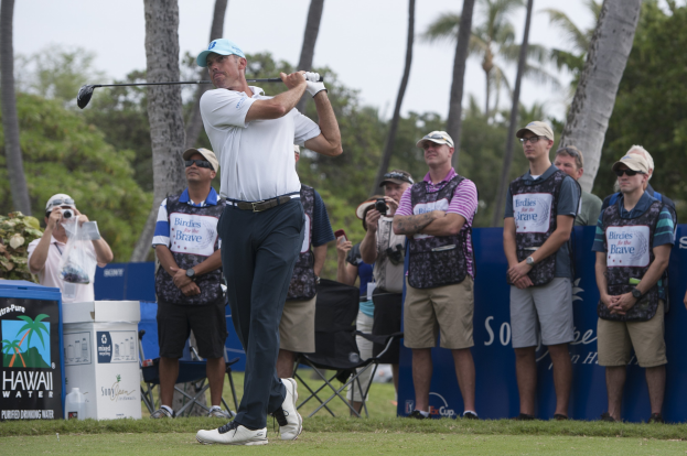 Ein Mann in einem weißen T-Shirt, schwarzen Hosen und einer blauen Mütze schwingt einen Golfschläger auf einem grünen Feld, mit Zuschauern und Kameras hinter ihm, vor einem Hintergrund aus Bäumen und einem klaren blauen Himmel.