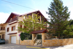 Eine Villa mit einem vor ihr geparkten Auto, das Fenster, Geländer, Säulen und ein Dach hat und von einem Tor, einem Schild, einem Straßenpfahl, Bäumen und einer Wand umgeben ist.