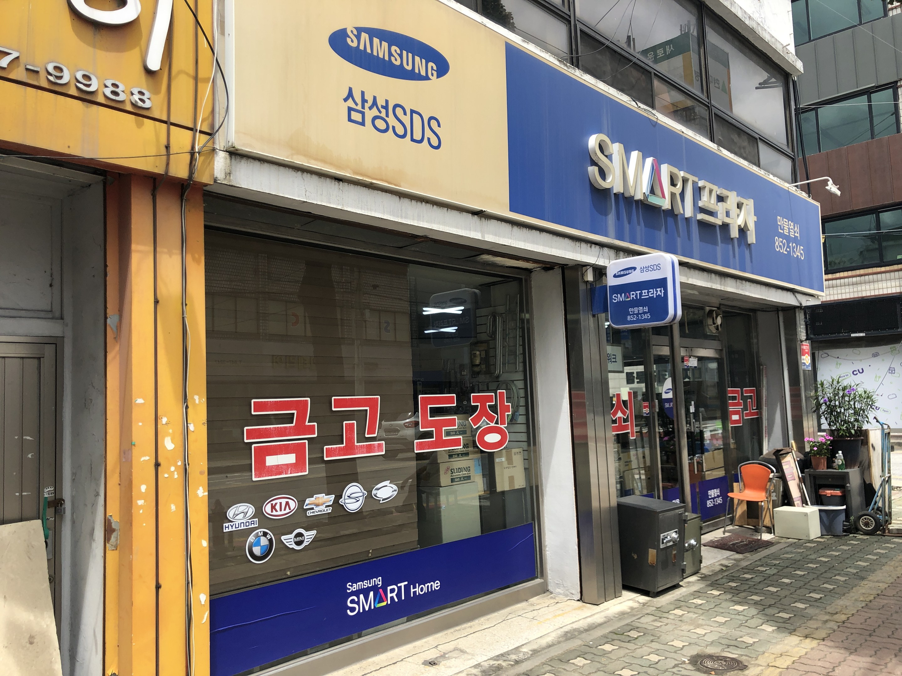 Außenansicht eines Gebäudes mit einer "Samsung Smart Home"-Plakette, umgeben von glaswandigen Gebäuden mit Namensschildern, Mülltonnen, Stühlen, Pflanzen und einem sichtbaren Himmel im Hintergrund.