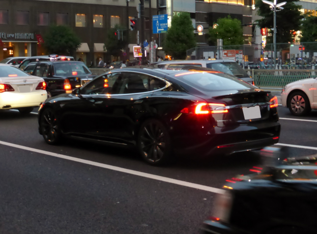 Schwarzer Tesla Model S fährt auf einer Stadtstraße bei Nacht mit anderen Fahrzeugen, Gebäuden, Bäumen, Laternen, Verkehrszeichen, Geländern und Fußgängern im Hintergrund.