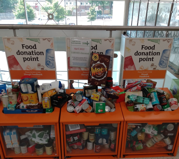 Orange Schübe mit Lebensmittelspenden, darunter Flaschen, Dosen und Kisten, mit Informationsschildern, durch Glasfenster mit Außenfahrzeugen, Bäumen, Gebäuden und Himmel.