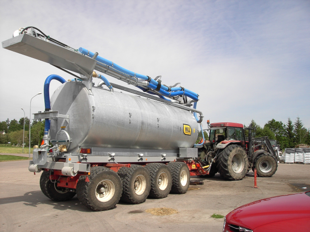 Ein großer Tanklastzug steht auf einem Parkplatz neben einem roten Auto, mit Bäumen, Polen, Lampen und einem klaren blauen Himmel im Hintergrund.