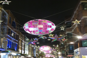 Eine festlich geschmückte Stadtstraße bei Nacht mit zahlreichen Lichtern und Dekorationen, darunter eine große Diskokugel, die von einem Gebäude hängt.