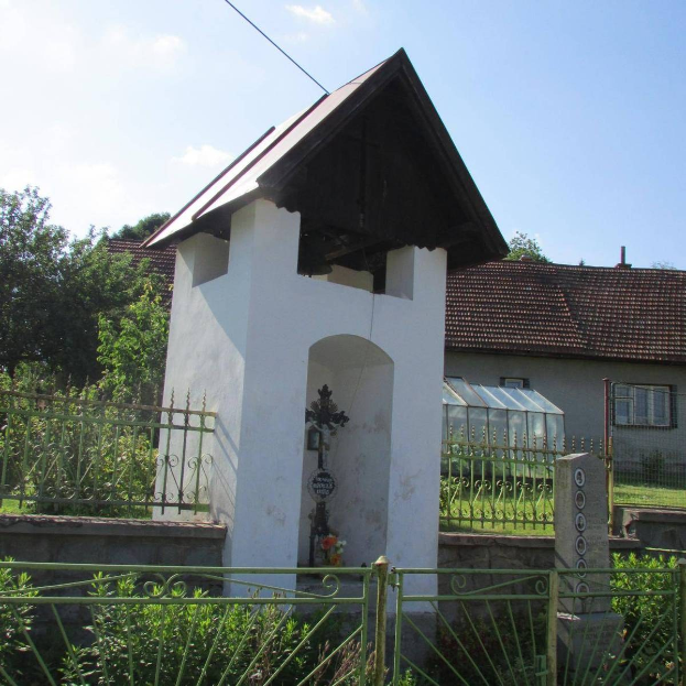 Kleine weiße Kirche mit einem Kreuz darauf, umgeben von einem Metallzaun, nahe einem Weg und Häusern unter einem bewölkten Himmel.