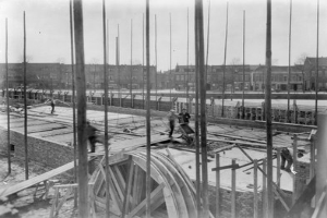 Schwarz-weiß-Foto einer Baustelle mit Arbeitern, Holzpfählen, einem Rad im Vordergrund und Gebäuden, Bäumen und einem klaren Himmel im Hintergrund.