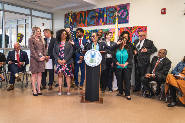 Gruppe von Menschen vor einem Rednerpult mit Mikrofon und einer Tafel mit Text, einige sitzen auf Stühlen, andere stehen, mit Flaggen, einer Tafel, einem Feuerlöscher und Deckenlampen an der Rückwand.