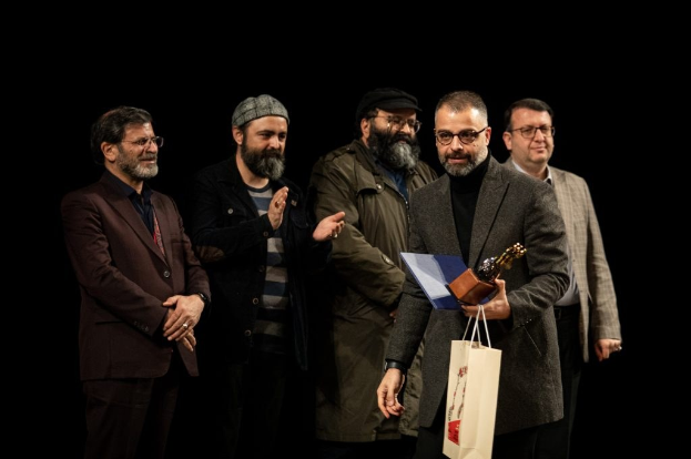 Eine Gruppe von Männern auf einer Bühne, einer hält eine Tasche und eine Trophäe, mit einem dunklen Hintergrund, der die Eröffnungsnacht des Berliner Filmfestivals andeutet.