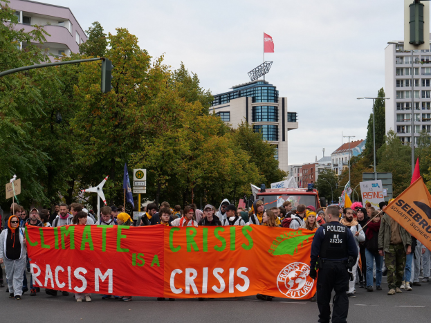 Eine Gruppe von Menschen marschiert auf einer von Bäumen gesäumten Straße, die ein Banner mit der Aufschrift 'Klimakrise ist eine Krise' trägt, mit Gebäuden und einem klaren Himmel im Hintergrund.