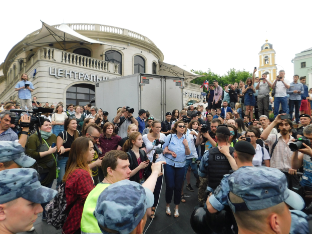 Große Gruppe von Menschen protestiert vor einem Gebäude, einige halten Kameras und Handys, mit einem geparkten Fahrzeug, Bäumen und einem klaren blauen Himmel im Hintergrund.
