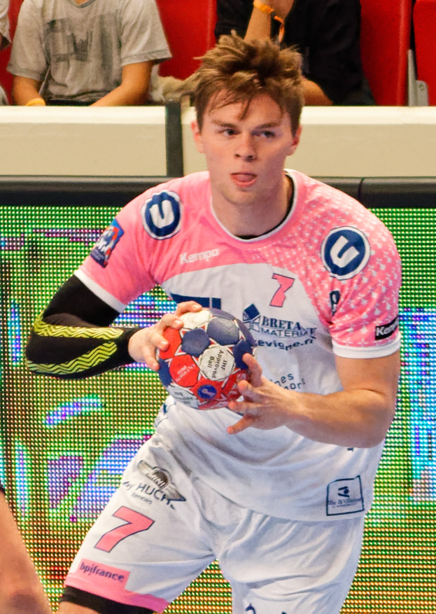 Ein Mann in einem pinken Hemd, der einen Fußball hält, mit einer anderen Person Hand auf der linken Seite, während im Hintergrund Menschen auf Stühlen sitzen.