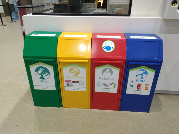 Vier bunte Mülltonnen in einer Reihe auf dem Boden mit einer Theke, Monitor und anderen Gegenständen im Hintergrund und einer Person links daneben.