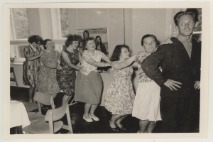 Eine Gruppe von Menschen, die in einem Raum im 1950er-Stil tanzen, mit Stühlen, einem Tisch, einem Foto Rahmen an der Wand und Fenstern.