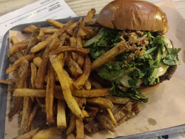 Ein Tablett mit einem Hamburger, der mit Salat, Tomate und Zwiebeln garniert ist und goldenen, knusprigen Pommes frites, das auf einem Tisch mit einem Blatt Papier daneben steht.