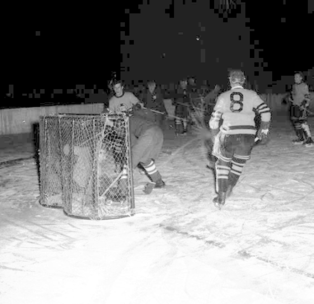 Schwarzes und weißes Bild von Männern, die Hockey auf einem Eisplatz spielen, mit einem Netz im Vordergrund und einer Wand im Hintergrund.