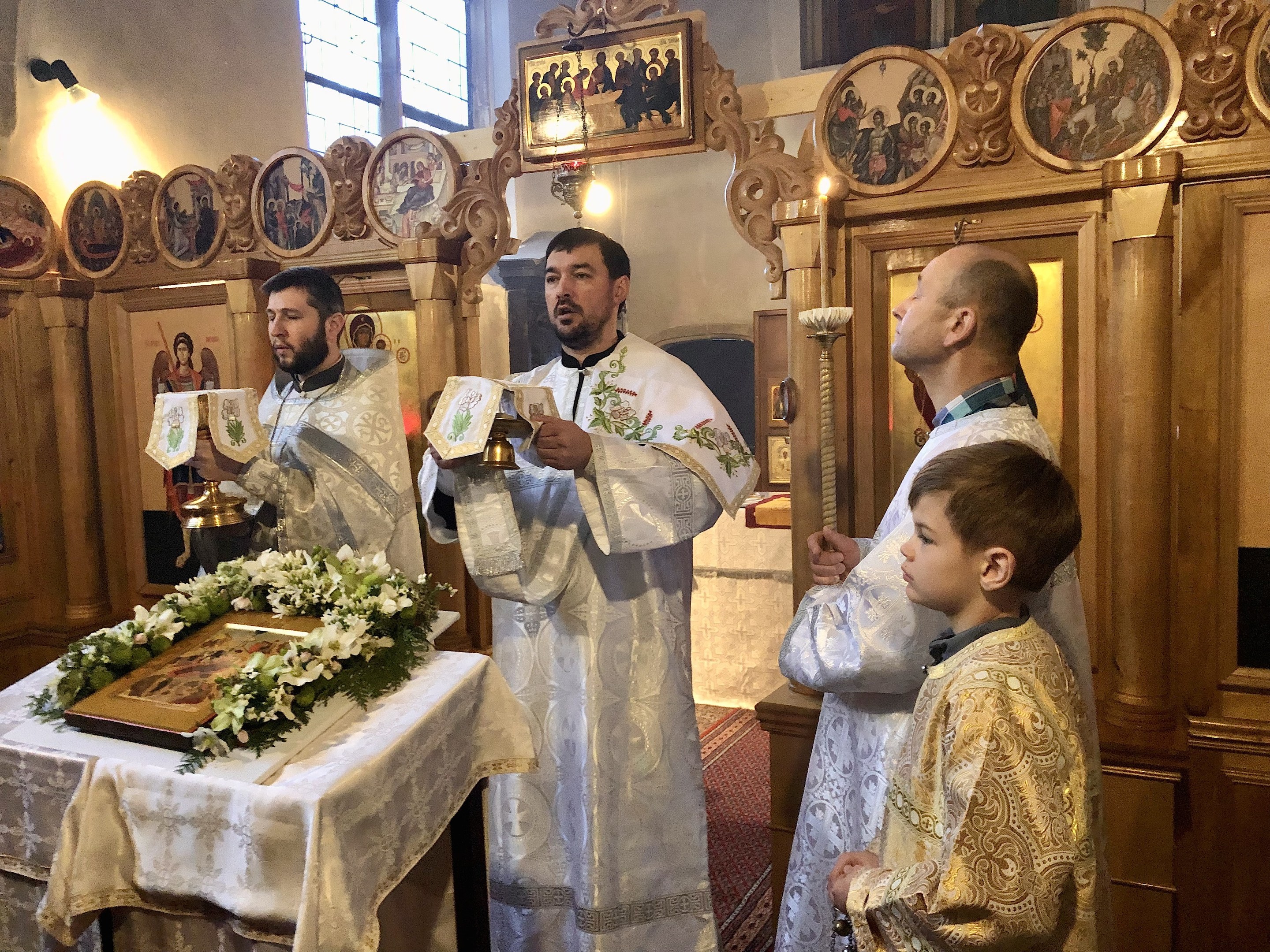 Eine Gruppe von Menschen steht um einen Tisch in einer Kirche, hält Bücher in den Händen, mit einem Tuch und einem Blumenstrauß auf dem Tisch und Foto Rahmen, Lampen, ein Fenster und ein hölzernes Objekt im Hintergrund.