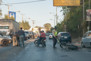 Eine Gruppe von Menschen steht um ein verunglücktes Motorrad auf der Straße mit mehreren Fahrzeugen, darunter ein Lastwagen, und einer Hintergrundkulisse aus Bäumen, Polen, Lampen, Schildern und Himmel.