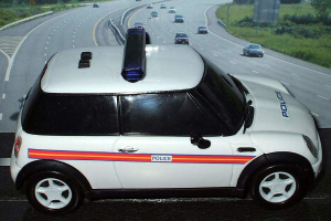 Ein weißer Polizeiwagen steht vor einer belebten Straße mit fahrenden Fahrzeugen, umgeben von Gras und Bäumen in einer ländlichen Umgebung.