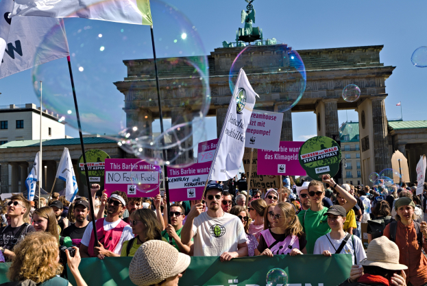 Eine Menschenmenge in Hüten und Brillen steht vor dem Brandenburger Tor in Berlin, Deutschland, einige halten Fahnen und Schilder, darunter eines im Vordergrund, während Gebäude, ein Tor mit einer Statue und ein Himmel mit Seifenblasen im Hintergrund zu sehen sind.