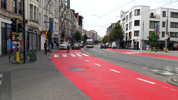 Roter Fahrradweg neben einer Stadtstraße mit fahrenden Fahrzeugen, Fußgängern auf dem Gehweg, Gebäuden mit Fenstern, Bäumen und einem klaren blauen Himmel im Hintergrund.