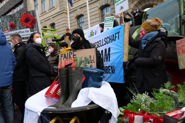 Eine Gruppe von Menschen mit Masken hält Protestschilder vor einem Lastwagen, mit einem Tisch mit Gegenständen, Topfpflanzen und Gebäuden im Hintergrund.