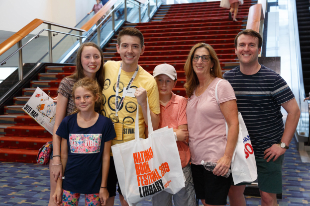 Gruppe von Menschen, die vor einer Rolltreppe an der Nationalen Buchmesse 2019 lächeln und Taschen halten, mit Treppen und Geländern im Hintergrund.