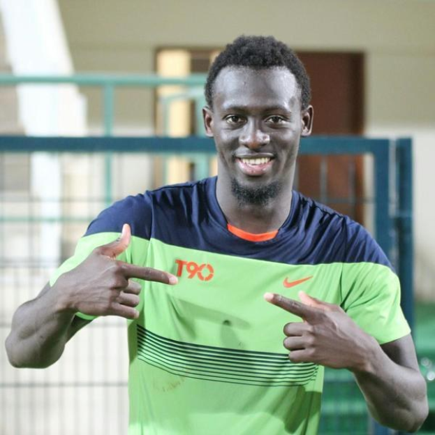 Ein Mann in einem grün-blauen Hemd, der lachend in die Kamera zeigt und im Hintergrund eine undeutliche Reling, Wand und Tür zu sehen sind.