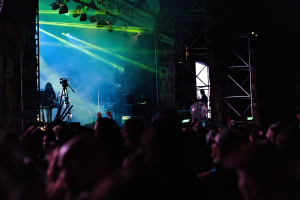 Eine Menge schaut Performern auf einer hell erleuchteten Bühne bei einem Konzert zu, mit Instrumenten, Mikrofonen und einem Banner.
