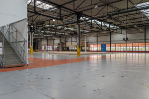 Ein großes industrielles Lagerhaus mit zahlreichen Fenstern, Metallstützen, einer Treppe mit Geländer, einem Schild mit Text, einem Dach mit Deckenleuchten und zusätzlichen Stützen.