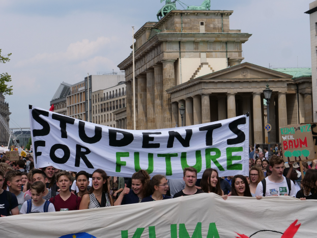 Gruppe von Schülern marschiert in Berlin mit einer bunt bemalten "Students for Future"-Schriftzug-Tafel an Gebäuden, Bäumen und Himmel vorbei.