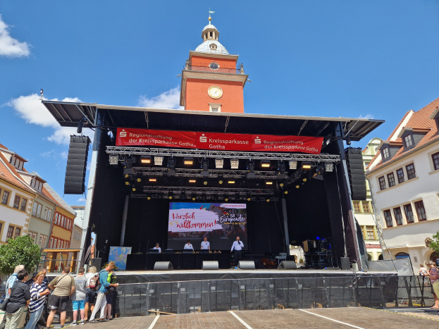 Gruppe von Menschen auf einer Bühne vor einem Uhrturm auf dem Kreissparkasse Gotha Musikfestival, mit Gebäuden, Bäumen und einem bewölkten Himmel im Hintergrund.