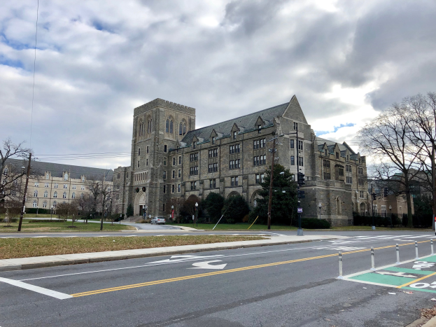 Großes Gebäude mit zahlreichen Fenstern, als Campus der Universität Pennsylvania bezeichnet, umgeben von Bäumen, Straßenlaternen, Ampeln und Fahrzeugen unter einem bewölkten Himmel.