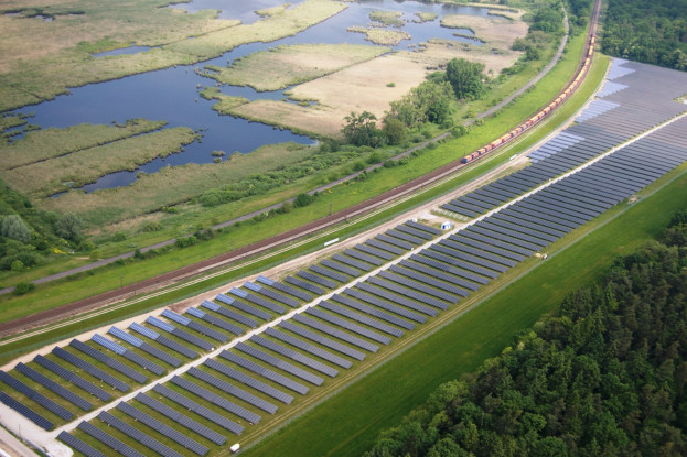 Luftaufnahme eines Solarparks mit Solarmodulen in einem Feld, umgeben von Bäumen, Gras, Pflanzen und Wasser, mit einem Zug auf einer nahen Bahnstrecke.