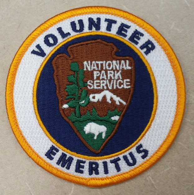 Runder Aufnäher mit blauem Rand und weißem Hintergrund, der in weißer Schrift den Text 'Volunteer National Park Service Emeritus' in der Mitte eines gelben Kreises zeigt.