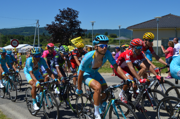 Gruppe von Radfahrern mit Helmen, Brillen, Handschuhen und Schuhen auf einer grasgesäumten Straße mit Zelten, Polen, Laternen, Schildern, Bäumen, Gebäuden, Hügeln und einem klaren blauen Himmel im Hintergrund.