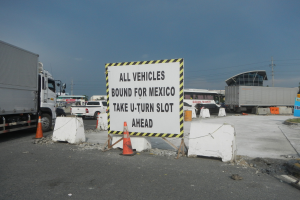 Lastwagen auf einem Parkplatz neben einem Schild mit der Aufschrift "Alle Fahrzeuge nach Mexiko" geparkt, mit verstreuten Verkehrskegeln, Brettern und Hintergrundgebäuden, Bäumen, Pfählen und einem klaren blauen Himmel.