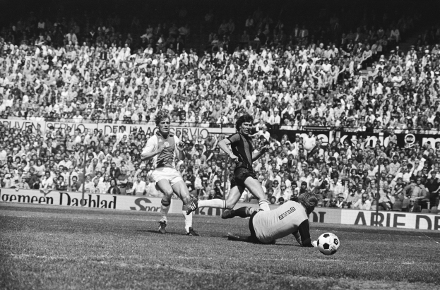 Schwarzes Foto von Männern, die Fussball auf einem Feld mit Zuschauern im Hintergrund und einer Bundesliga 1978-79 Ajax 1-1 Hannover 96 Fahne spielen.
