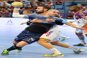 Eine Gruppe von Männern spielt Handball in einem Stadion, wobei ein Spieler den Ball hält und Zuschauer im Hintergrund sitzen.