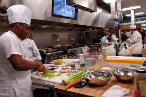 Eine Gruppe von Köchen in einer Küche, die auf einem Schneidbrett Lebensmittel zubereiten, wobei ein Koch in der foreground einen weißen Kittel und eine Mütze trägt und ein Messer hält und von Utensilien, Schüsseln, Gemüse, einem Mobiltelefon, Flaschen und anderen Gegenständen umgeben ist, mit einem Fernseher und Deckenlampen im Hintergrund.