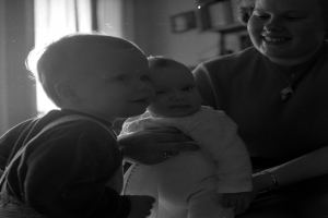 Eine Frau hält ein Baby mit einem warmen Lächeln, mit zwei anderen Kindern neben ihr, in einem gemütlichen Raum mit Rahmen und einem Fenster im Hintergrund.