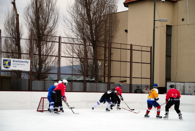 Menschen, die Eis-hockey auf einem Eisplatz mit Gebäuden, Bäumen, einer Straßenlaterne, einem Namensschild und Zäunen im Hintergrund unter einem klaren Himmel spielen.