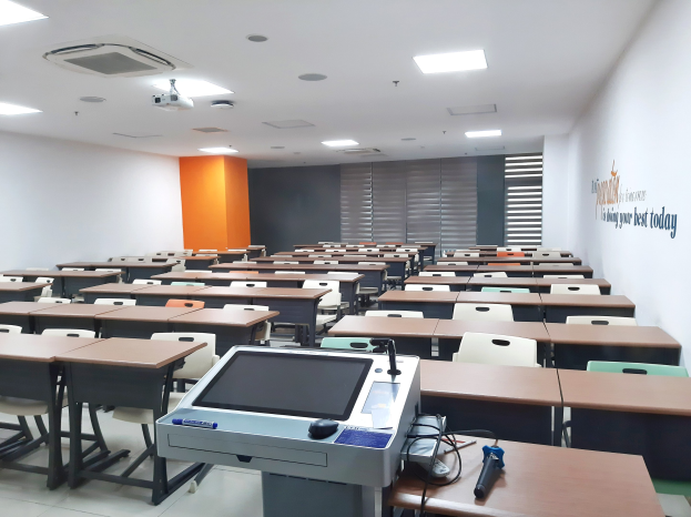 Ein Klassenzimmer mit Tischen und Stühlen in Reihen, ein deckenmontierter Projektor und Gegenstände auf den Tischen, mit Text an der rechten Wand, der darauf hinweist, dass es sich um eine Privatschule in Singapur handelt.