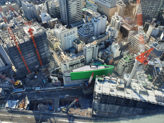 Luftaufnahme einer Stadt mit hohen Gebäuden, Straßen, Fahrzeugen, Bäumen und einer großen grünen Baustelle in der Mitte.