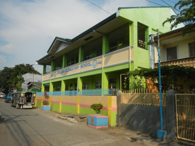Schule mit grüner und gelber Fassade, Fahrzeugen auf der Straße, Topfpflanzen, einem Zaun, einem Tor, einem Straßenpfosten, Bäumen, Stromleitungen und einem bewölkten Himmel.